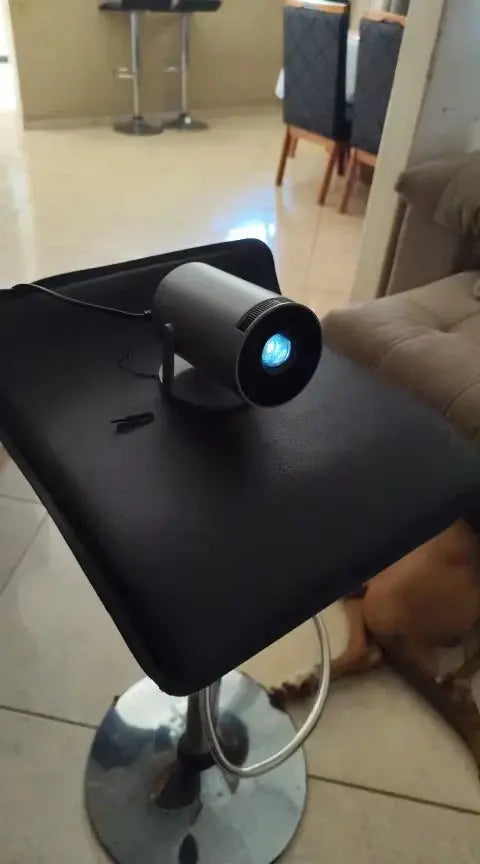 Compact mini projector placed on a black leather stool, projecting a bright image in a cozy indoor setting